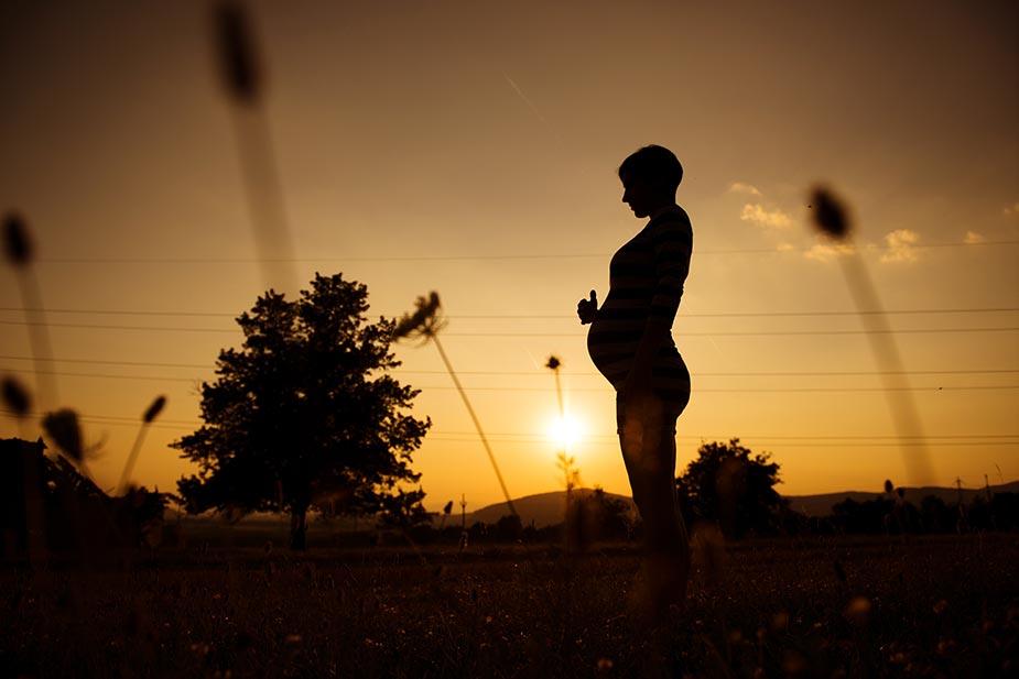 Silueta trudnice iz profila koja drži ruke na stomaku. U pozadini zalazak sunca.