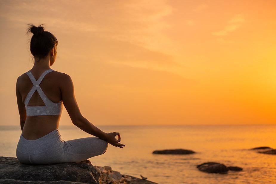 Žena sedi na obali mora u lotos položaju. Sunce zalazi i more je mirno.