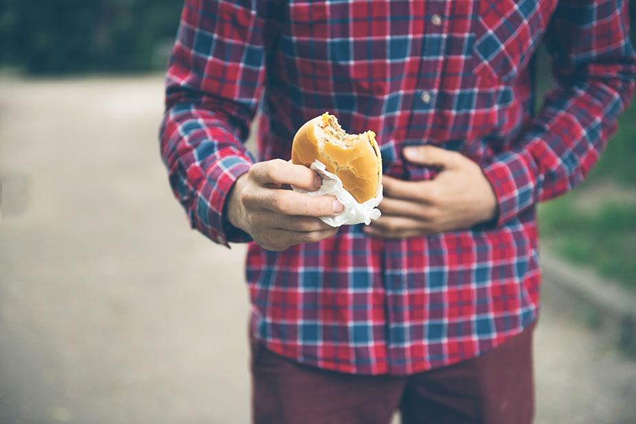 Mladić kome se ne vidi gornji deo tela jednom rukom drži lepinju sa hamburgerom a drugom stomak